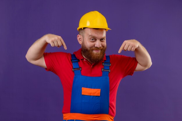 Young bearded builder man in construction uniform and safety helmet smiling confident pointing to himself with index fingers over purple background