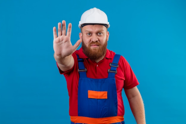 Giovane barbuto uomo costruttore in uniforme da costruzione e casco di sicurezza in piedi con la mano aperta facendo segno di stop cercando dispiaciuto su sfondo blu