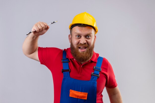 Young bearded builder man in construction uniform and safety helmet swinging a wrench with aggressive expression