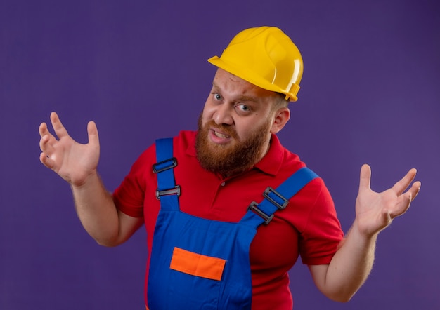 Giovane uomo barbuto costruttore in uniforme da costruzione e casco di sicurezza con espressione aggressiva frustrato con le braccia alzate su sfondo viola