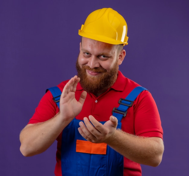 Young bearded builder man in construction uniform and safety helmet with confident smile on face applauding over purple background