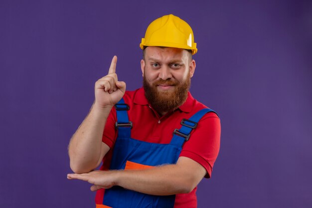 紫色の背景の上に指を上向きに懐疑的な表情で建設制服と安全ヘルメットの若いひげを生やしたビルダーの男