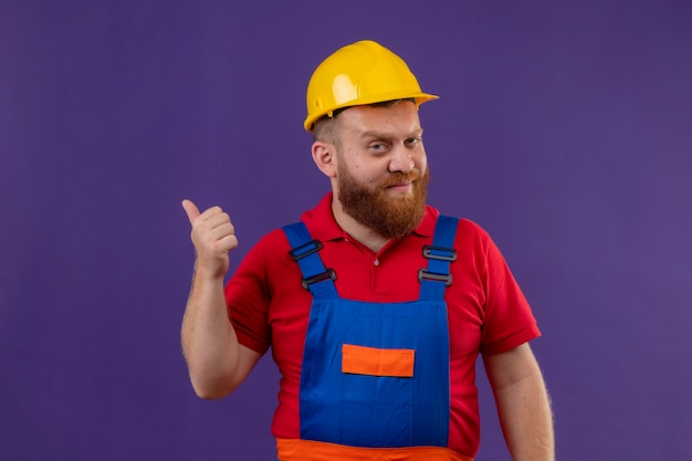 Young bearded builder man in construction uniform and safety helmet with skeptic expression pointing with thumb to something behind over purple background