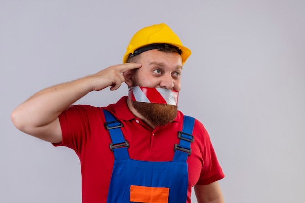 Giovane uomo barbuto costruttore in uniforme da costruzione e casco di sicurezza con nastro adesivo sulla bocca che guarda da parte confuso indicando la tempia