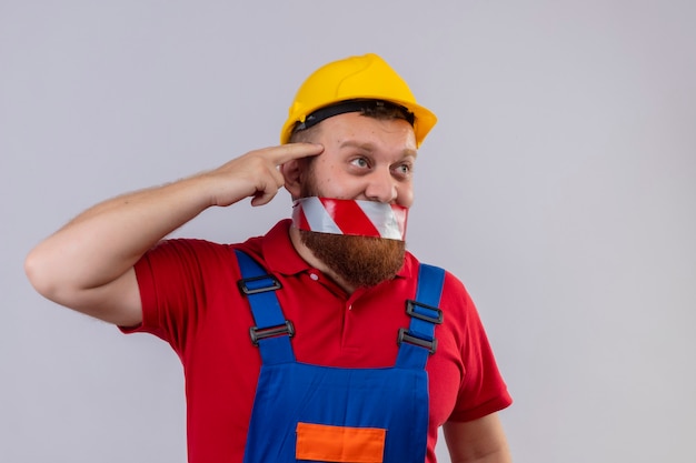 Young bearded builder man in construction uniform and safety helmet with tape over his mouth looking aside confused pointing his temple