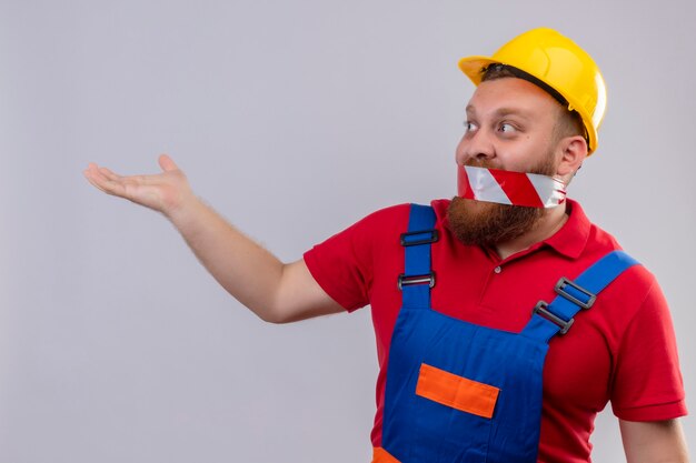 Young bearded builder man in construction uniform and safety helmet with tape over his mouth looking aside surprised with raised hand