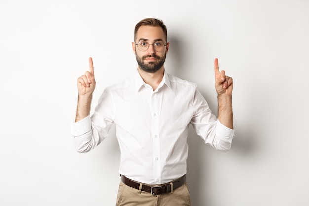 Young bearded businessman pointing fingers up, showing promo offer, standing  