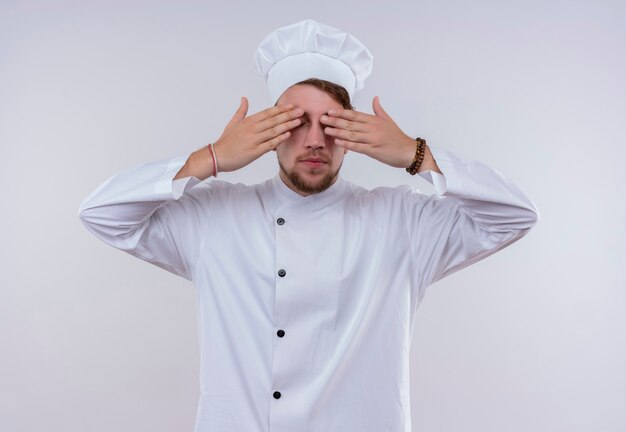 A young bearded chef man wearing white cooker uniform and hat covering his eyes with hands on a white wall