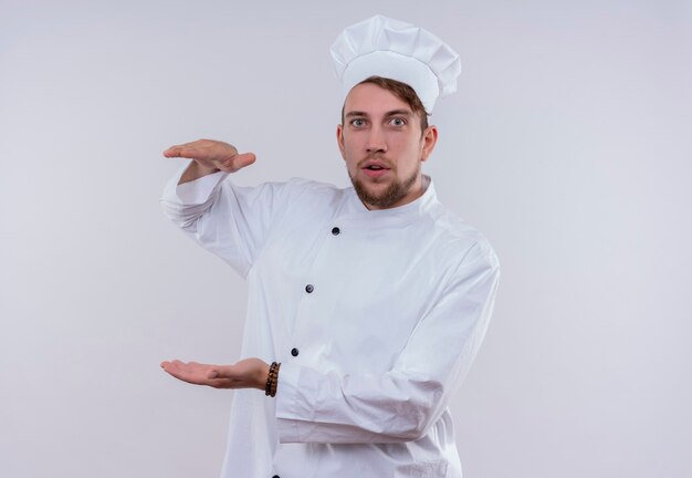 A young bearded chef man wearing white cooker uniform and hat gesturing with hands big and large size sign while looking on a white wall