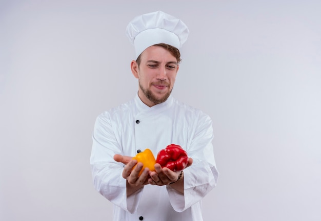 Молодой бородатый шеф-повар в белой униформе и шляпе смотрит на желтый и красный сладкий перец на руке на белой стене