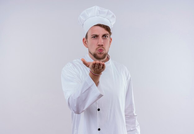 흰색 밥솥 유니폼과 모자를 쓰고 서서 흰 벽을 보는 동안 키스를 보내는 젊은 수염 난 요리사 남자