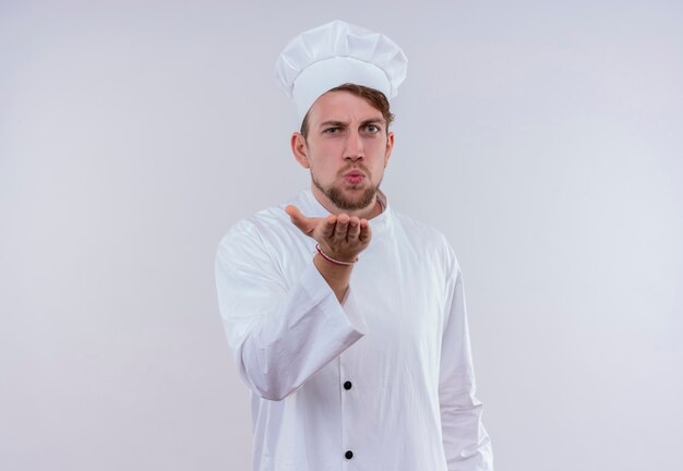 A young bearded chef man wearing white cooker uniform and hat sending a kiss while standing and looking on a white wall