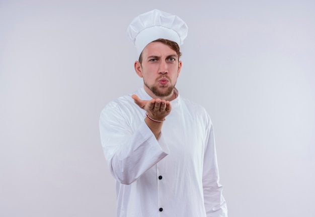 A young bearded chef man wearing white cooker uniform and hat sending a kiss while standing and looking on a white wall