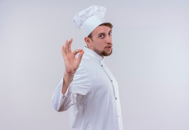 A young bearded chef man wearing white cooker uniform and hat showing ok sign with fingers while looking on a white wall