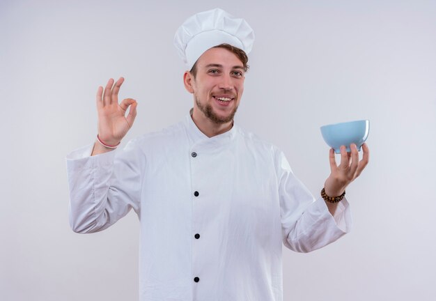 A young bearded chef man wearing white cooker uniform and hat showing tasty ok sign with fingers with blue bowl on his hand while looking on a white wall