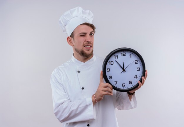 A young bearded chef man wearing white cooker uniform and hat showing wall clock and winking an eye while looking on a white wall