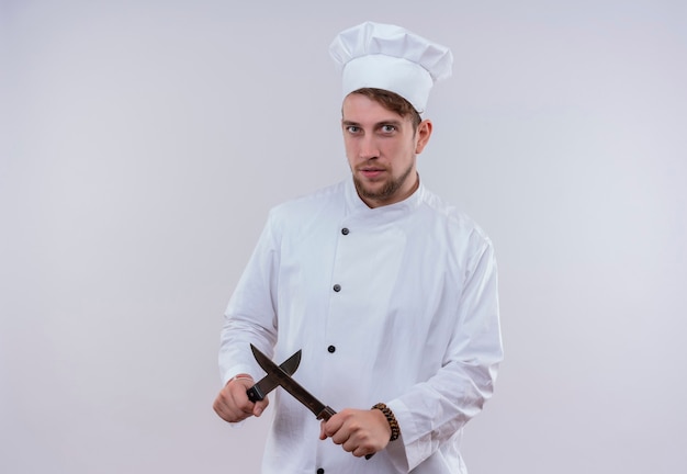 A young bearded chef man wearing white cooker uniform and hat showing x sign with knife while looking on a white wall