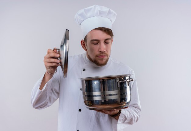 A young bearded chef man wearing white cooker uniform and hat smelling a cooking pan on a white wall