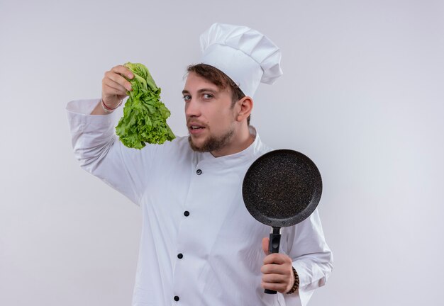 Молодой бородатый шеф-повар в белой форме держит сковороду и зеленый салат, глядя на белую стену
