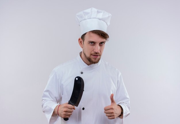 흰색 제복을 입은 젊은 수염 난 요리사 남자 고기 식칼을 잡고 흰 벽을 보면서 엄지 손가락을 보여주는