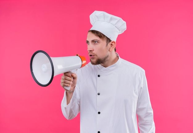 A young bearded chef man in white uniform holding megaphone and speaking through it on a pink wall