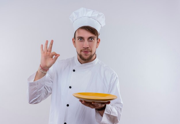 A young bearded chef man in white uniform holding yellow plate and showing ok sign gesture while looking on a white wall