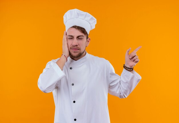 A young bearded chef man in white uniform pointing up with index finger while holding hand on face on an orange wall