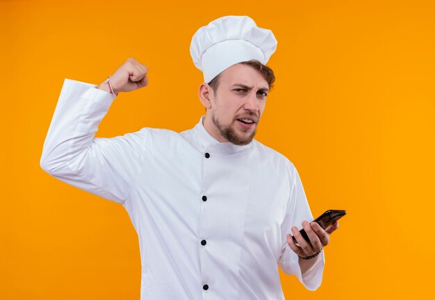 A young bearded chef man in white uniform raising clenched fist and holding mobile phone while looking on an orange wall