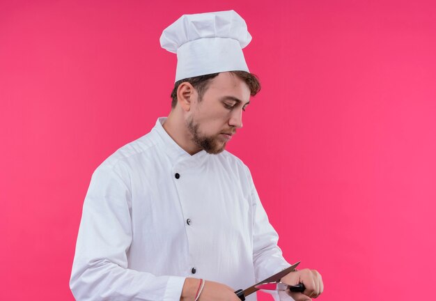 분홍색 벽에 서있는 동안 칼을 날카롭게하는 흰색 제복을 입은 젊은 수염 난 요리사 남자