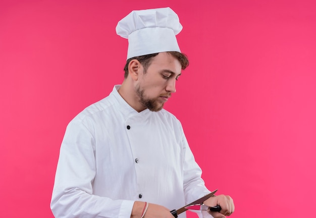 Молодой бородатый шеф-повар в белой униформе точит нож, стоя на розовой стене