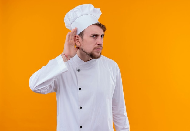A young bearded chef man in white uniform trying to listen to something on an orange wall