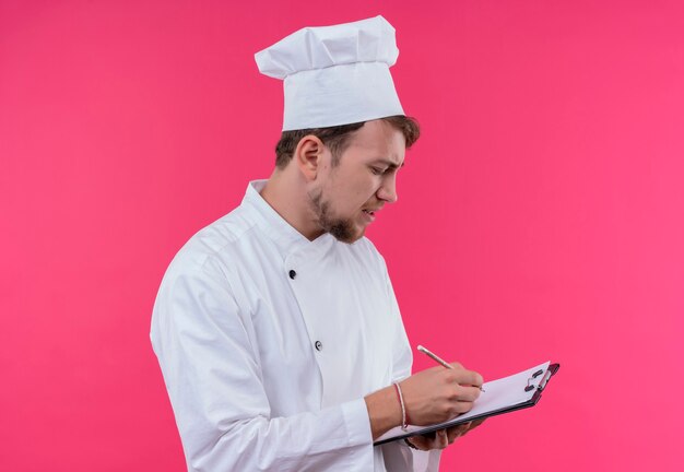 A young bearded chef man in white uniform writing something on notepad folder while standing on a pink wall