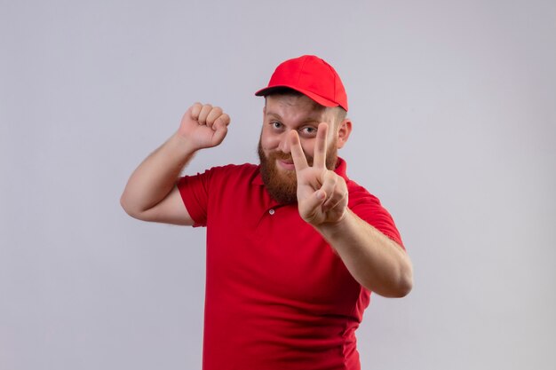 Молодой бородатый курьер в красной форме и сжимая кулак в кепке, показывая знак победы улыбается