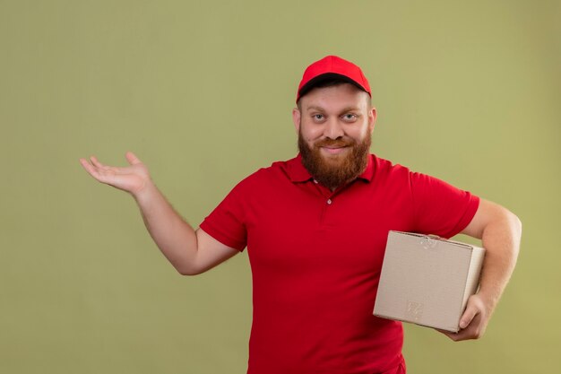 Young bearded delivery man in red uniform and cap holding cardboard box looking at camera with confident smile presenting with arm of his hand