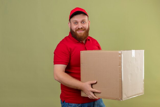 Young bearded delivery man in red uniform and cap holding cardboard box looking at camera with confident smile