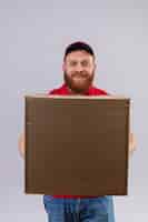 Free photo young bearded delivery man in red uniform and cap holding cardboard box looking at camera with confident smile