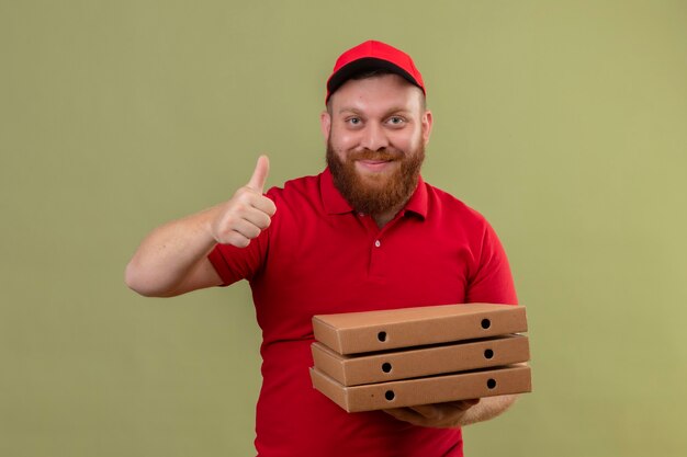 Young bearded delivery man in red uniform and cap holding stack of pizza boxes looking at camera smiling showing thumbs up