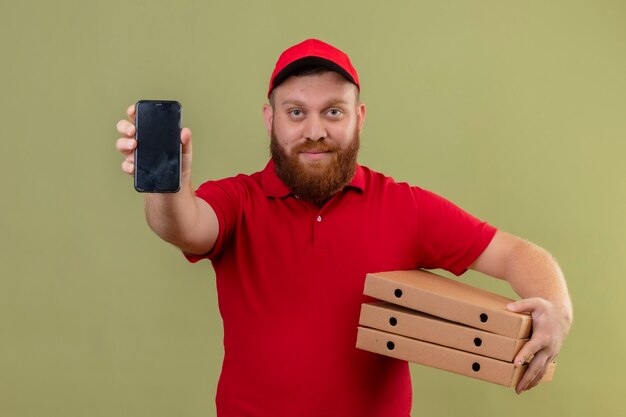 Young bearded delivery man in red uniform and cap holding stack of pizza boxes showing smartphone to camera looking confident