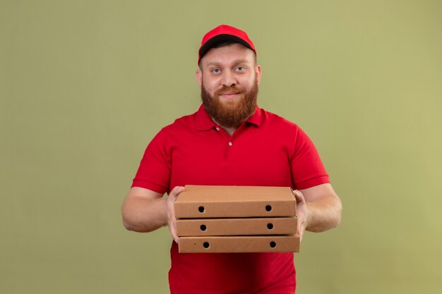 Young bearded delivery man in red uniform and cap holding stack of pizza boxes smiling confident