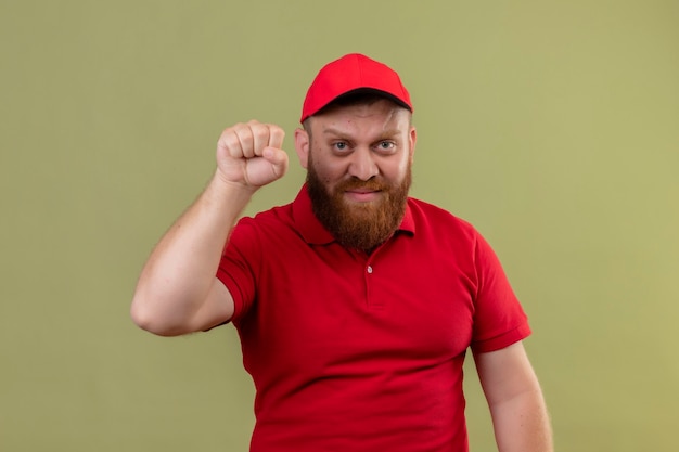 Молодой бородатый курьер в красной форме и кепке, поднимающий кулак как победитель, смотрит в камеру с серьезным лицом