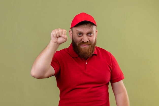 Young bearded delivery man in red uniform and cap raising fist as a winner, looking at camera with serious face