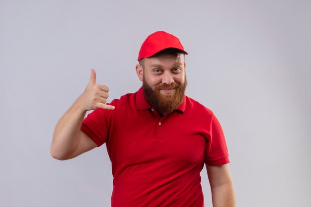 Young bearded delivery man in red uniform and cap smiling making cal me gesture with hand