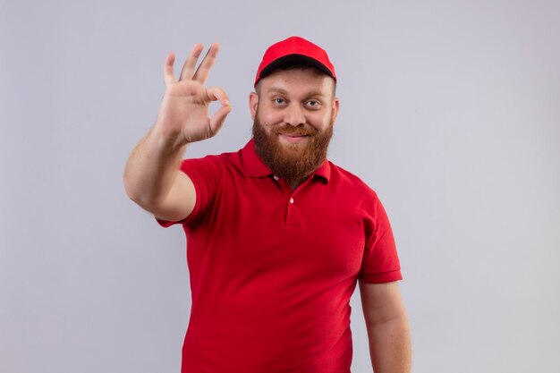 Young bearded delivery man in red uniform and cap smiling positive and happy doing ok sign