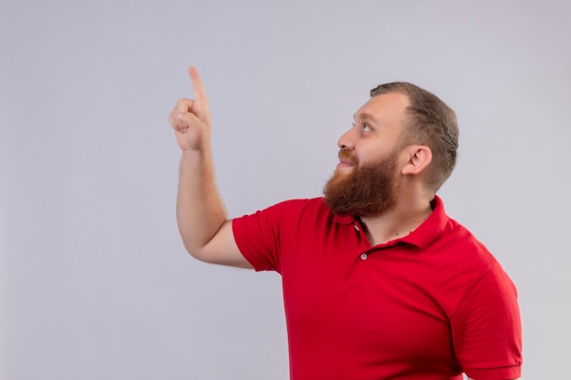 Free photo young bearded delivery man in red uniform looking aside pointing up with index finger