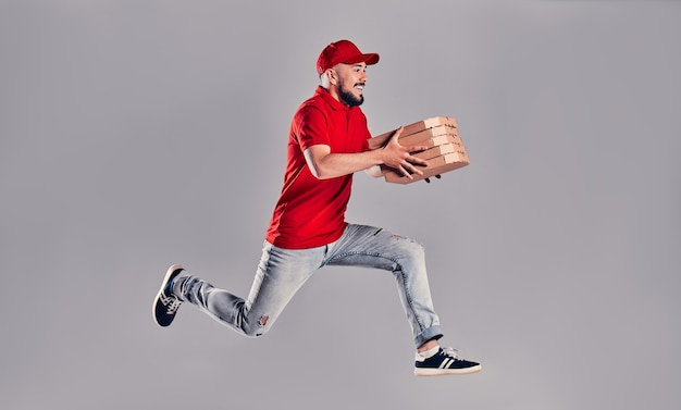 Young bearded delivery man in a red uniform runs in a jump flight holds boxes with pizza isolated on gray background. fast home delivery of pizza. Premium Photo