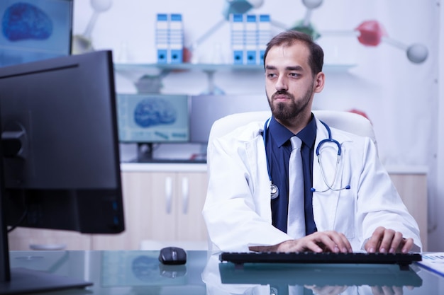 Giovane medico barbuto con entrambe le mani sulla tastiera che digita la ricetta per un paziente. dottore concentrato sul suo lavoro in clinica.