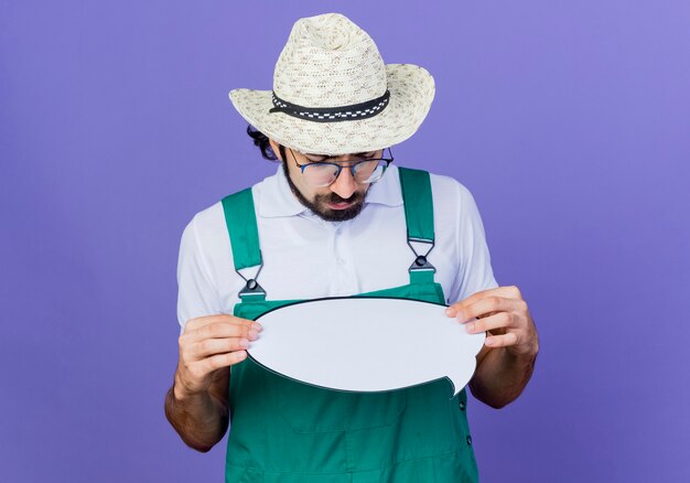 young bearded gardener man wearing jumpsuit and hat holding blank speech bubble sign looking at it with serious face standing over blue wall