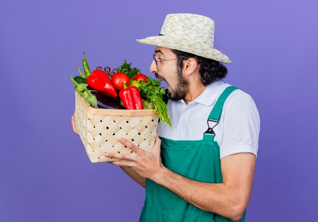 Young bearded gardener man wearing jumpsuit and hat holding crate full of vegetables biting tomato standing over blue wall