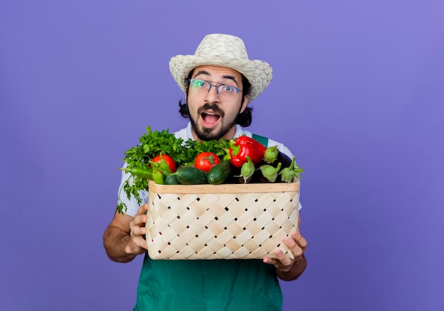 Foto gratuita giovane giardiniere barbuto uomo che indossa tuta e cappello tenendo la cassa piena di verdure guardando davanti felice ed eccitato in piedi sopra la parete blu