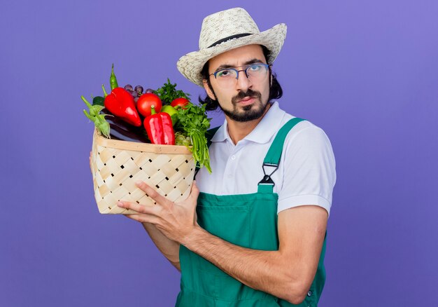 青い壁の上に立っている真面目な顔で正面を見て野菜でいっぱいの木枠を保持しているジャンプスーツと帽子を身に着けている若いひげを生やした庭師の男